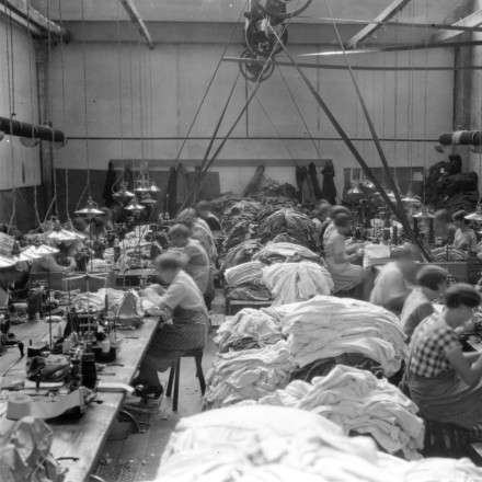 Schwarz Weiß Foto von Arbeiter*innen in einer alten Fabrik. Sie sitzen an Nähmaschinen, zwischen ihnen liegen Stoffberge.