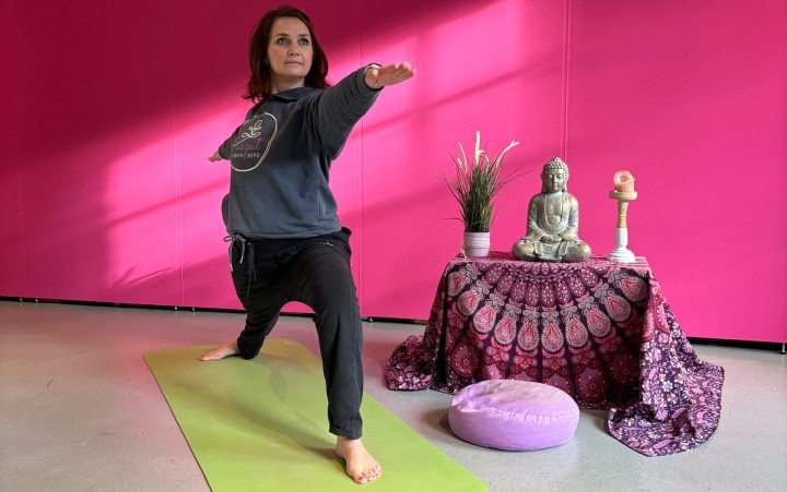 Person in bequemer Kleidung steht in Yogapose auf einer Matte vor einem kleinen Altar mit Buddha-Statue und Kerze vor einer pinkfarbenen Wand.