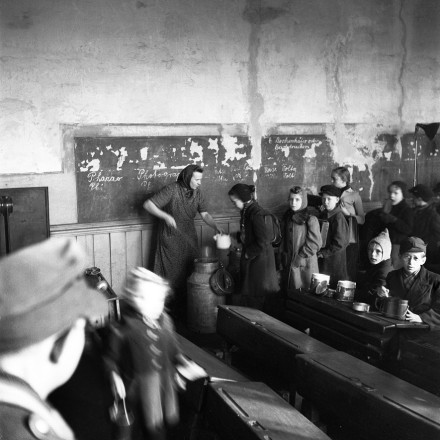 Historisches Foto eines Klassenzimmers. An der Wand steht eine Frau bei der Suppenausgabe. Kinder stehen in der Schlange, um Suppe von ihr zu bekommen. Andere Kinder sitzen mit Suppenschüsseln an den Schulbänken.