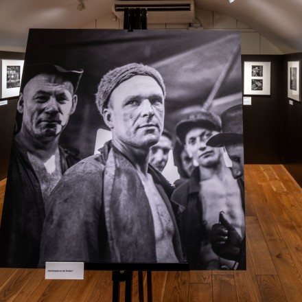 Foto aus der Ausstellung "Anne Winterer". Im Vordergrund ist ein historisches Foto von Männern, die als Kumpel im Ruhrgebiet gearbeitet haebn. Im Hintergrund sieht man weitere historische Fotos an der Wand hängen.