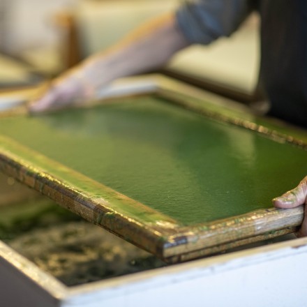 Eine Person hält einen Holzrahmen über ein Wasserbecken. Im Holzrahmen ist eine grüne und feuchte Papiermasse.