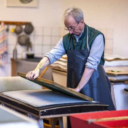 Ein Mann mit Schürze gautscht einen frischgeschöpften Papierbogen ab. 