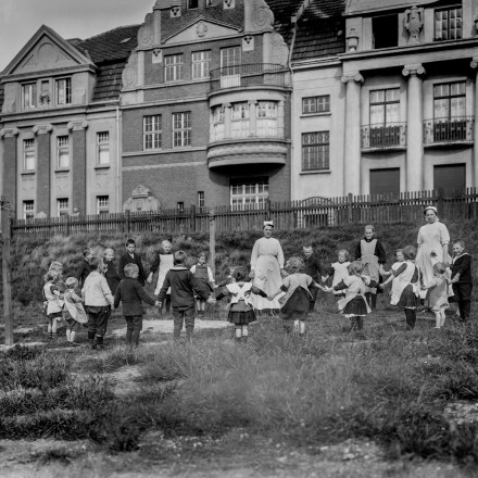 Historisches Foto von Kindern die sich an der Hand haltend mit einer erwachsenen Person im Kreis stehen. 