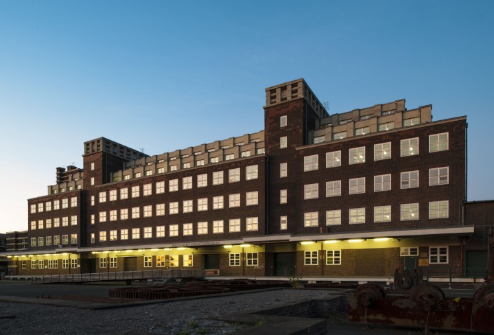 Gebäude mit vielen Fensterreihen (Peter-Behrens-Bau) in der Abenddämmerung. Am und im Gebäude sind Lichter eingeschaltet.