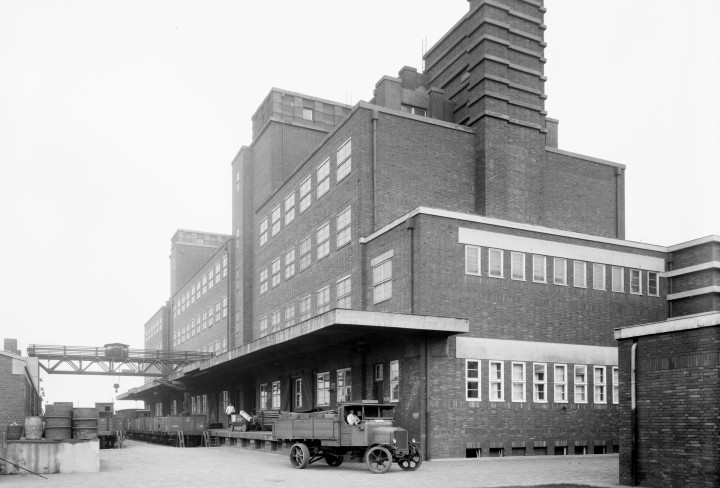 Schwarz-weiß Foto eines Gebäudes mit vielen Fenstern (Peter-Behrens-Bau). Vor dem Eingang steht ein Auto, das von Arbeitern be- und entladen wird.