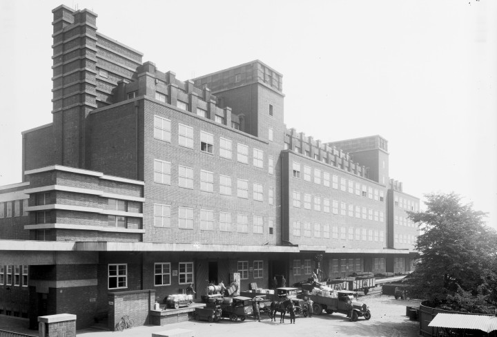 Schwarz-weiß Foto eines Gebäudes mit vielen Fenstern (Peter-Behrens-Bau). Vor dem Eingang stehen Autos und Kutschen, die be- und entladen werden.