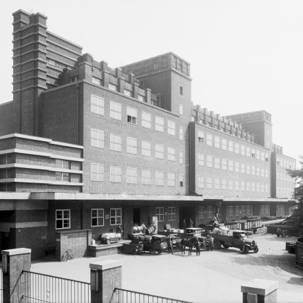 Der Peter-Behrens-Bau, ein langgestrecktes Gebäude mit sieben Stockwerken und einer Backsteinfassade. Im Vordergrund befindet sich eine Laderampe mit einem Fuhrwerk, zwei LKW, Eisenbahnwaggons und Elektrokarren. 