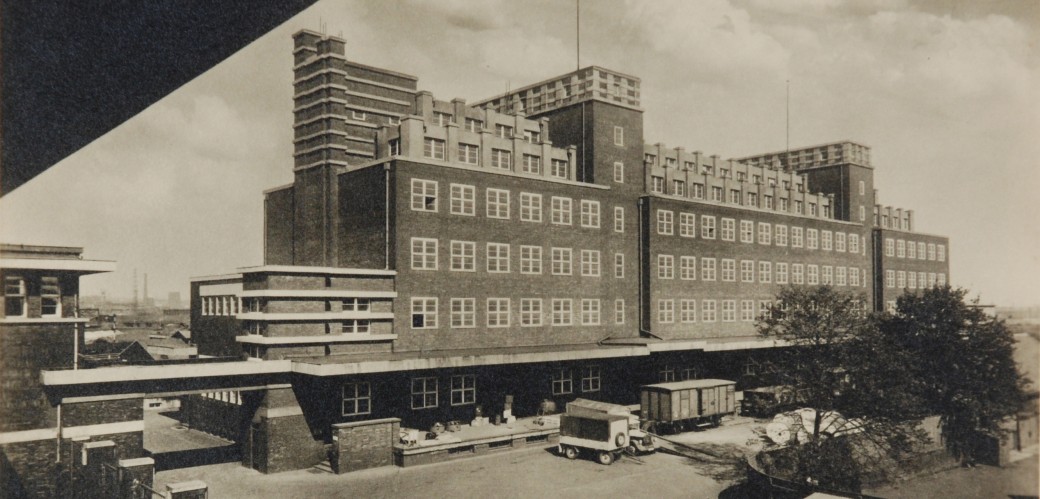 Black and white photograph of a large brick building. There are several cargo trailers and trees in front of the building. Black and white photograph of a large brick building. There are several cargo trailers and trees in front of the building.