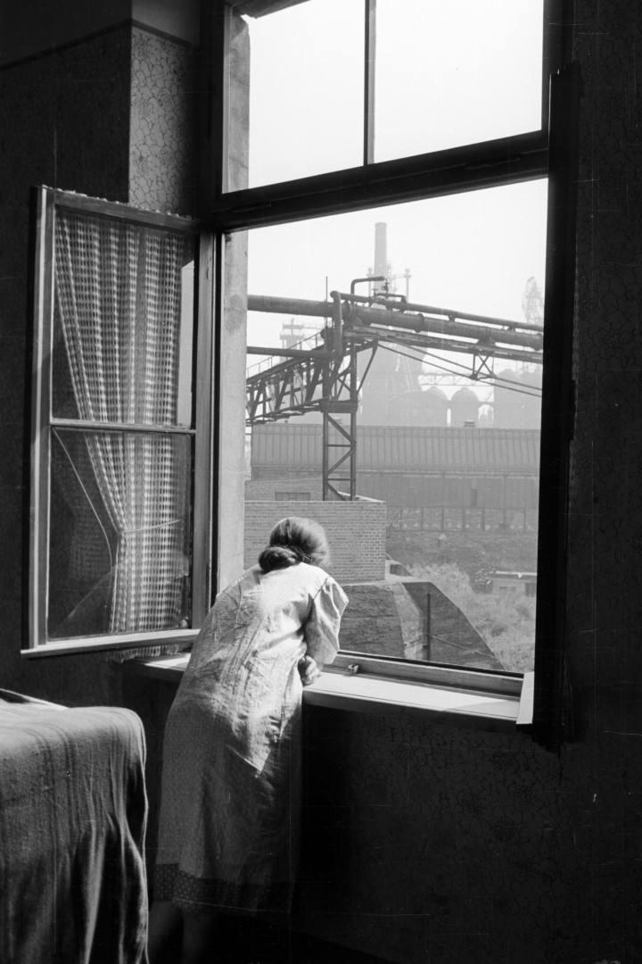 Schwarz-weiß Foto zeigt eine Frau die aus einen Fenster auf eine Industrielandschaft schaut