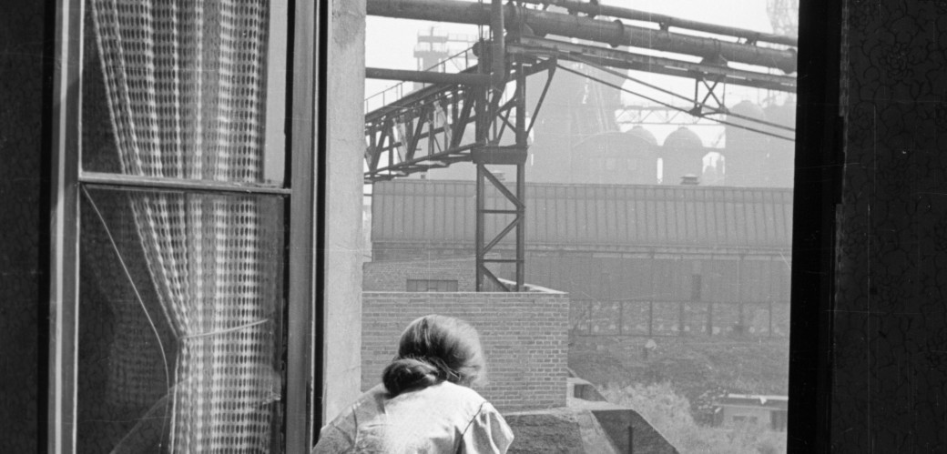 Schwarz-Weiß-Fotografie von 1952: Eine Frau steht vor einem geöffneten Fenster und schaut auf die unmittelbare Industrieanlage der August-Thyssen-Hütte in Duisburg