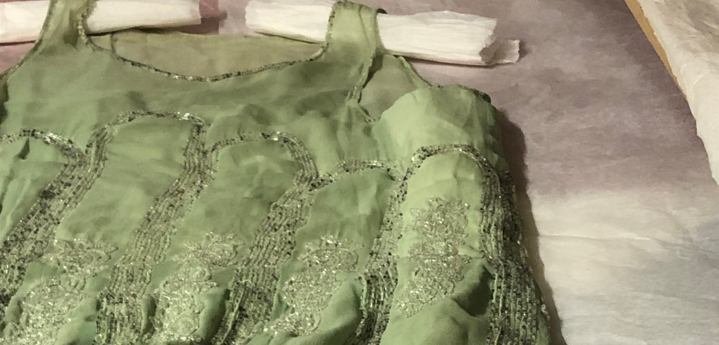 Auf einem ausziehbaren Holzregal im Textildepot liegt ein pastellgrünes Kleid aus feinem Chiffon. Es ist mit vielen Galsperlen besetzt.