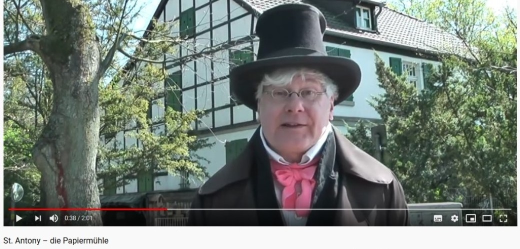 Screenshot eines Youtube Videos. In der Videoaufnahme befindet sich ein älterer Herr mit Zylinder und Gehrock bekleidet bei sonnigem Wetter vor einem weißen Fachwerkhaus.