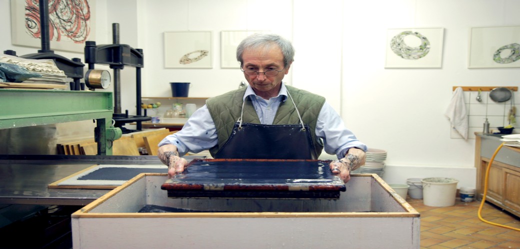 Person mit Schürze schöpft eine schwarze Flüssigkeit aus einem rechteckigen Behälter in einem Atelier mit Kunstwerken an der Wand