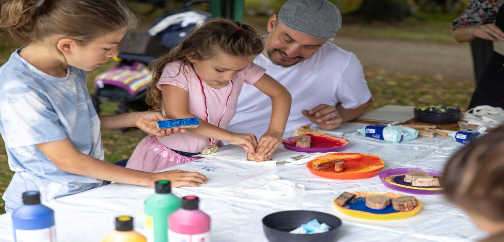 Kinder sitzen an einem Tisch und Stempeln mit bunten Farben auf ein weißes Tuch.