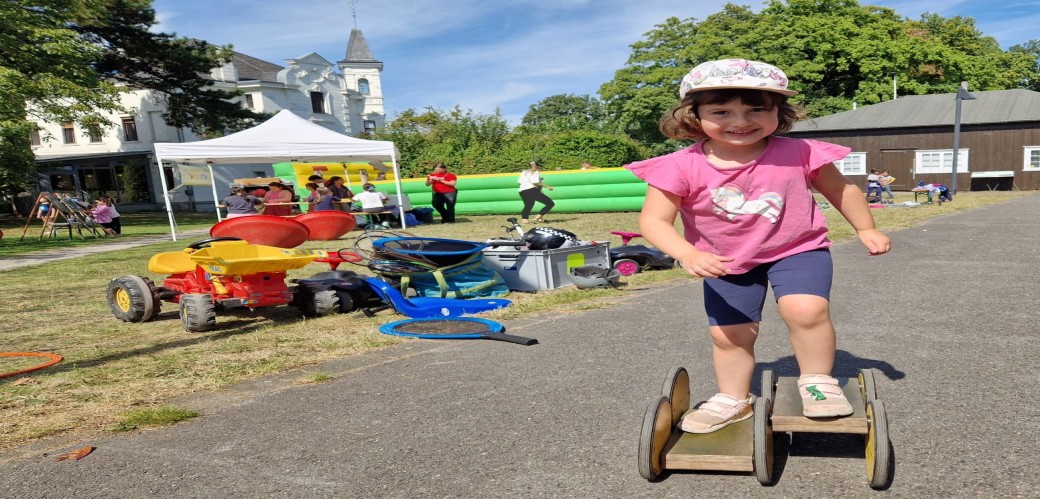 Kind mit rosa T-Shirt und Cap balanciert auf einem hölzernen Pedalo auf einem Weg in einem Park mit Spielgeräten und Menschen im Hintergrund.