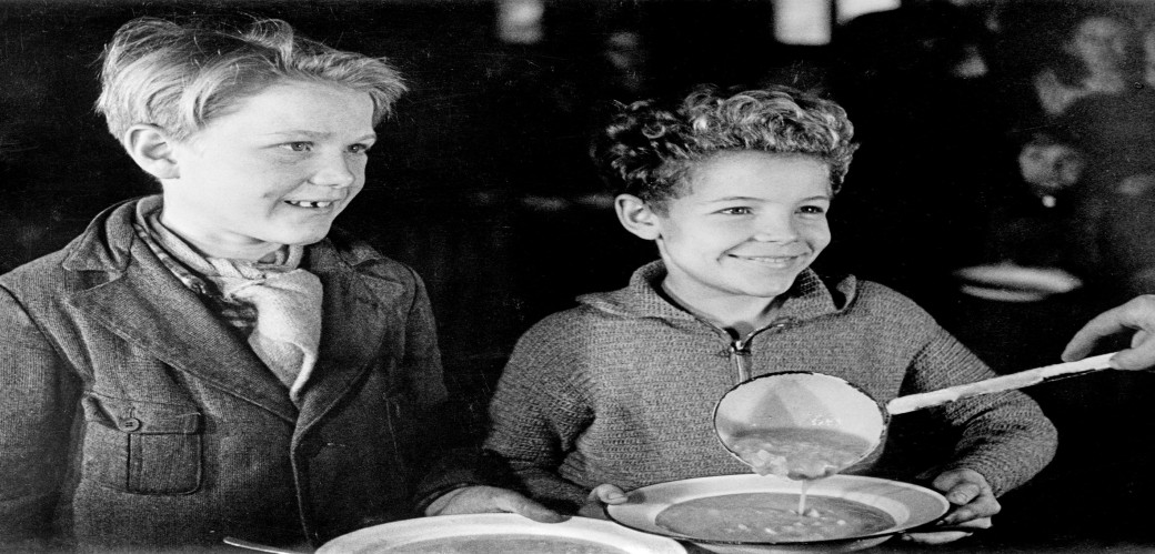 Schwarz/Weiß Aufnahme von zwei lächelnden Kindern mit Suppentellern, eine Hand schenkt mit einer Kelle Suppe ein. 