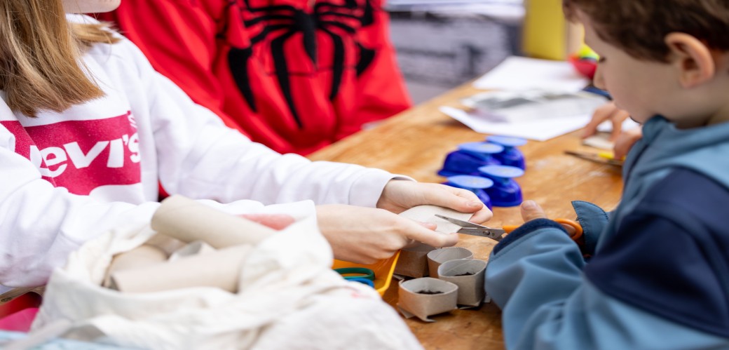 Kinder sitzen an einem Tisch und schneiden mit einer Schere Papierrollen aus Papprollen.