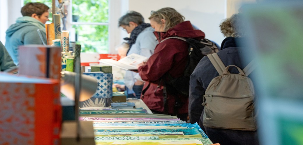 Mehrere Personen stehen an einem Tisch mit verschiedenen bunten Musterpapier, eine Person schaut interessiert in ein ausgestelltes Buch.