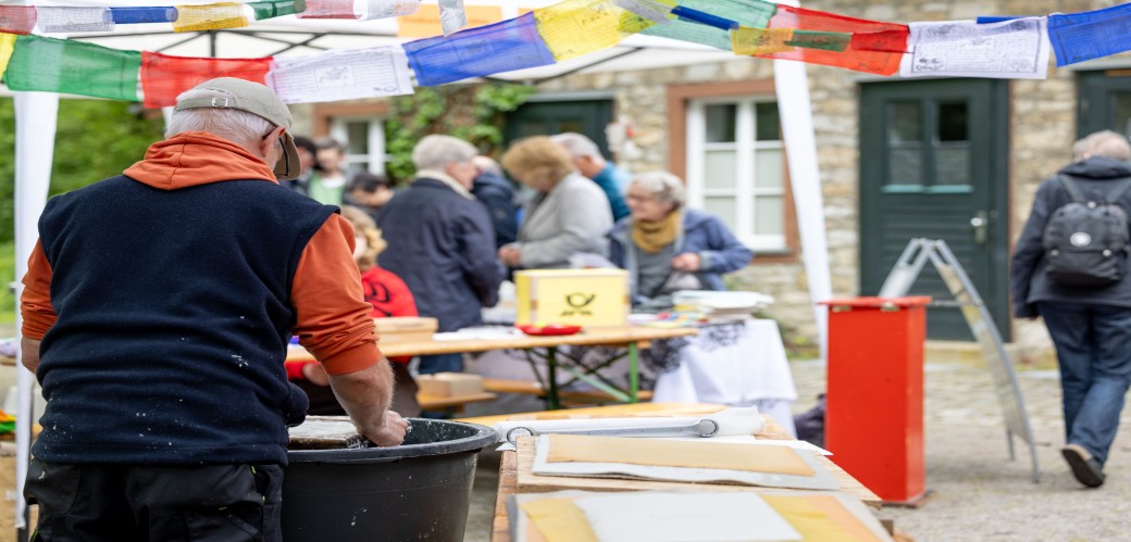 Mann mit rotem Pullover steht an einem Tisch mit Papierbögen, im Hintergrund mehrere Personen bei einem Markt im Freien
