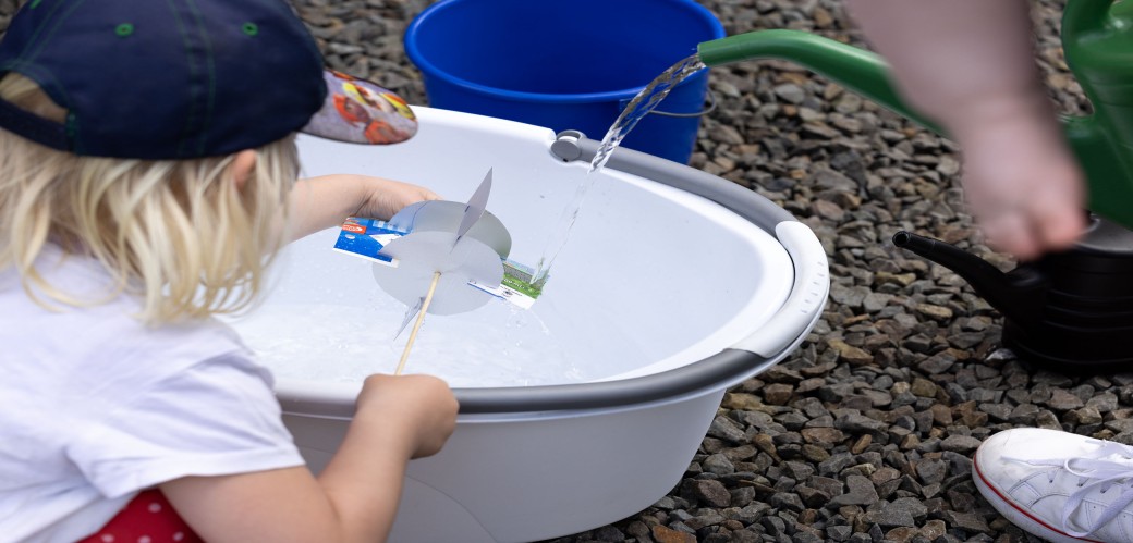 Kind hält selbstgebautes Wasserrad aus Papier über eine weiße Wanne, in die Wasser aus einem grünen Gartenschlauch fließt