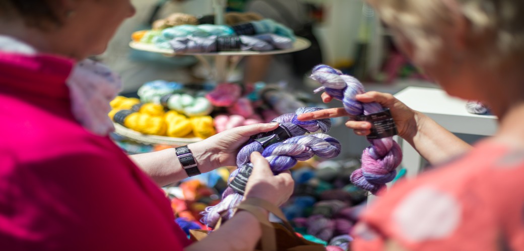  Zwei Frauen betrachten bunte Wollknäuel in einem Marktstand mit mehreren Regalen voller Wolle im Hintergrund. 