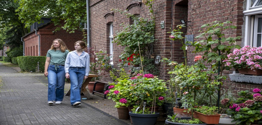 Zwei Frauen gehen auf einem Gehweg an einem Backsteinhaus mit vielen Blumentöpfen und Pflanzen vorbei