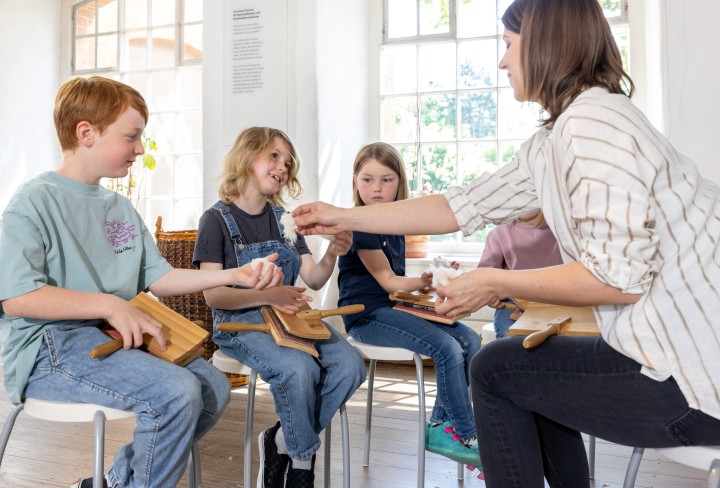 Drei Kinder und eine Frau sitzen in einem Stuhlkreis. Die Frau reicht den Kindern Baumwolle.
