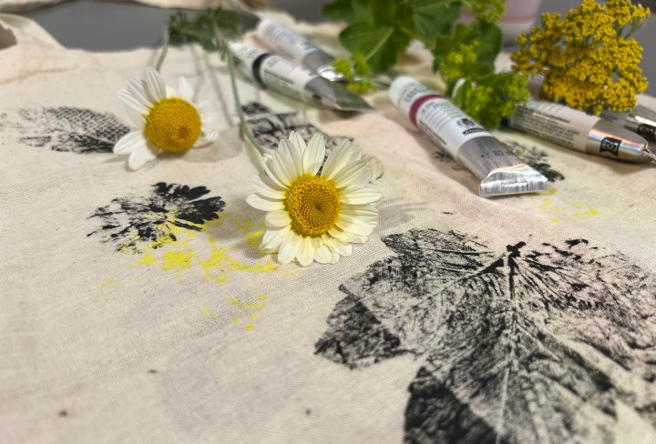 Auf dem Bild liegen zwei Gänseblümchen und einige Tuben mit Farbe auf einem weißen Stoffbeutel. Auf diesem sind Abdrücke von Blättern und Blüten zu sehen, die vermutlich mit Farbe gestaltet wurden. Im Hintergrund sind weitere Pflanzen und Farben erkennbar. 