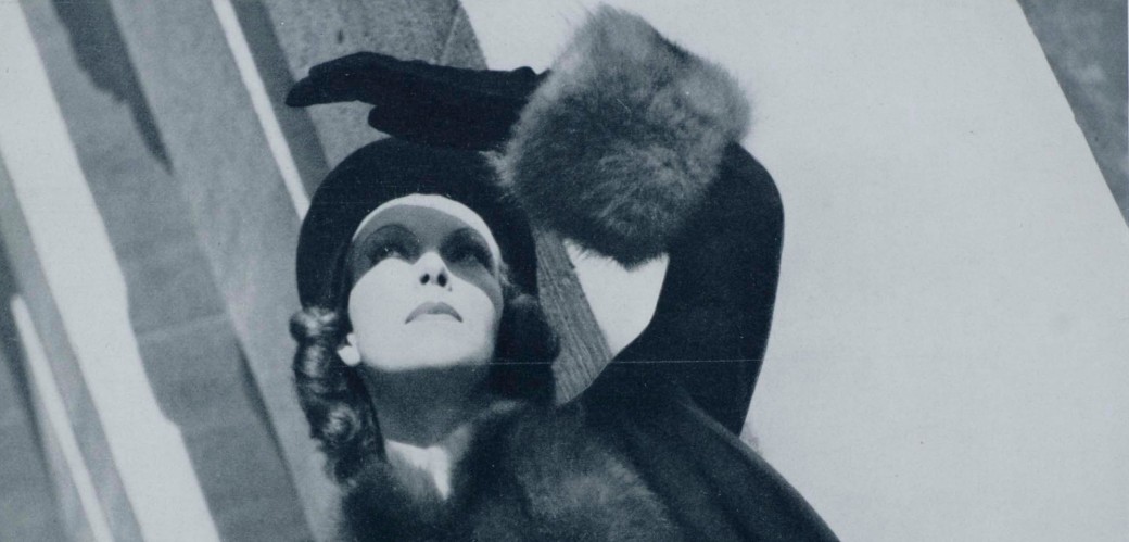 Eine Schwarz-Weiß-Fotografie zeigt eine Frau im kurzen Pelzmantel, die nach oben blickt.