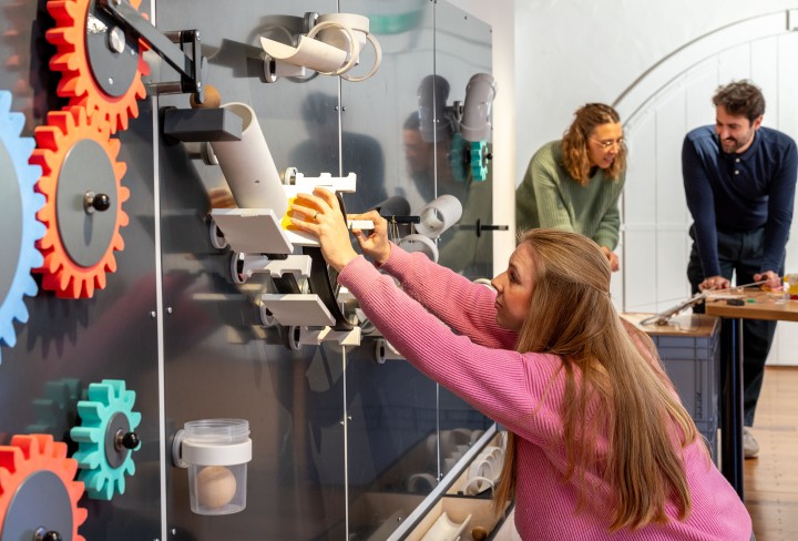 Eine Frau in einem rosa Pullover bedient eine interaktive Ausstellungswand mit Zahnrädern, Röhren und mechanischen Elementen. Im Hintergrund stehen zwei weitere Personen an einem Tisch. 