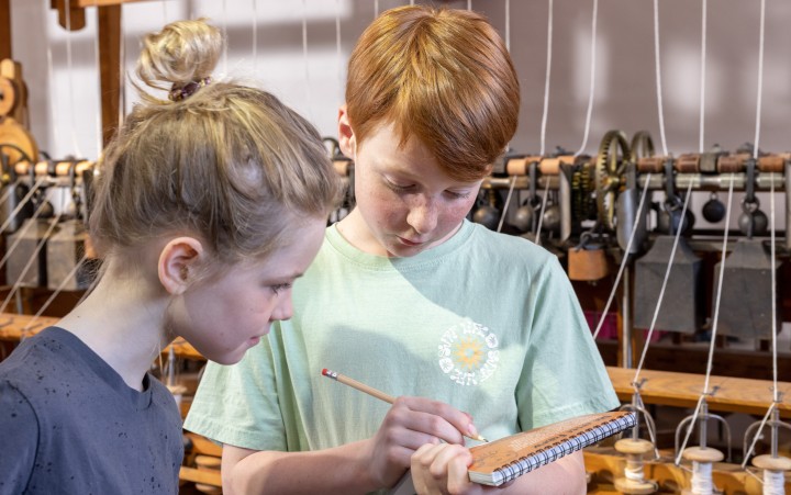 Zwei Kinder stehen vor einer historischen Webmaschine, eines hält ein Notizbuch und einen Bleistift in der Hand.
