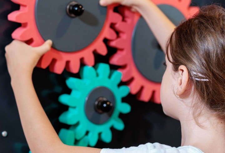Ein Mädchen dreht an drei bunten Zahnrädern, die an einer Wand hängen. 