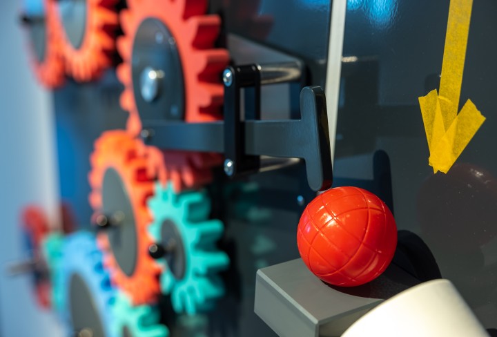 Eine rote Kugel liegt auf einer schwarzen kleinen Plattform. Im Hintergrund sind mehrere bunte Zahnräder an einer Wand. Auf die Kugel zeigt ein gelber Pfeil.