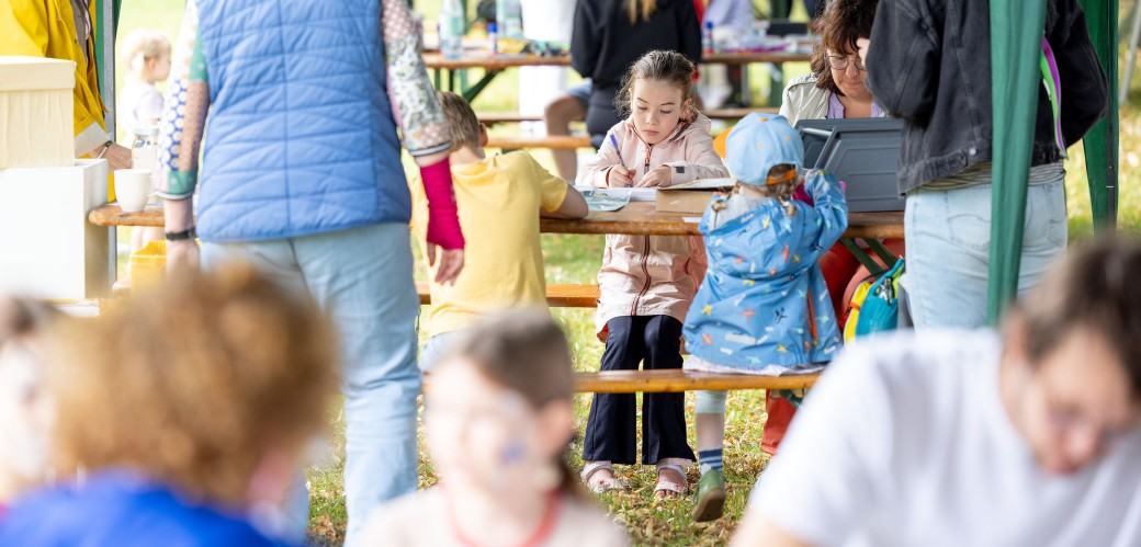 Viele Menschen unter einem Pavillion. Im Fokus sind Kinder, die an einem Tisch basteln und malen.