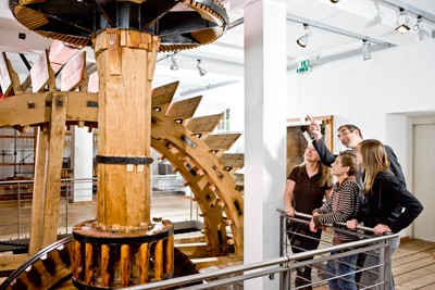 The power for the <br />“water frame” and the preparatory spinning machines was supplied by the water from the nearby Anger river. Up until the mid-19th century the factory was able to operate without steam power. Today when visitors enter the red brick building referred to as “Hohe Fabrik” (High Mill) they stand directly in front of a powerful wooden waterwheel which powers all the spinning machines in the museum via a transmission as was the case in the 18th century. <br />