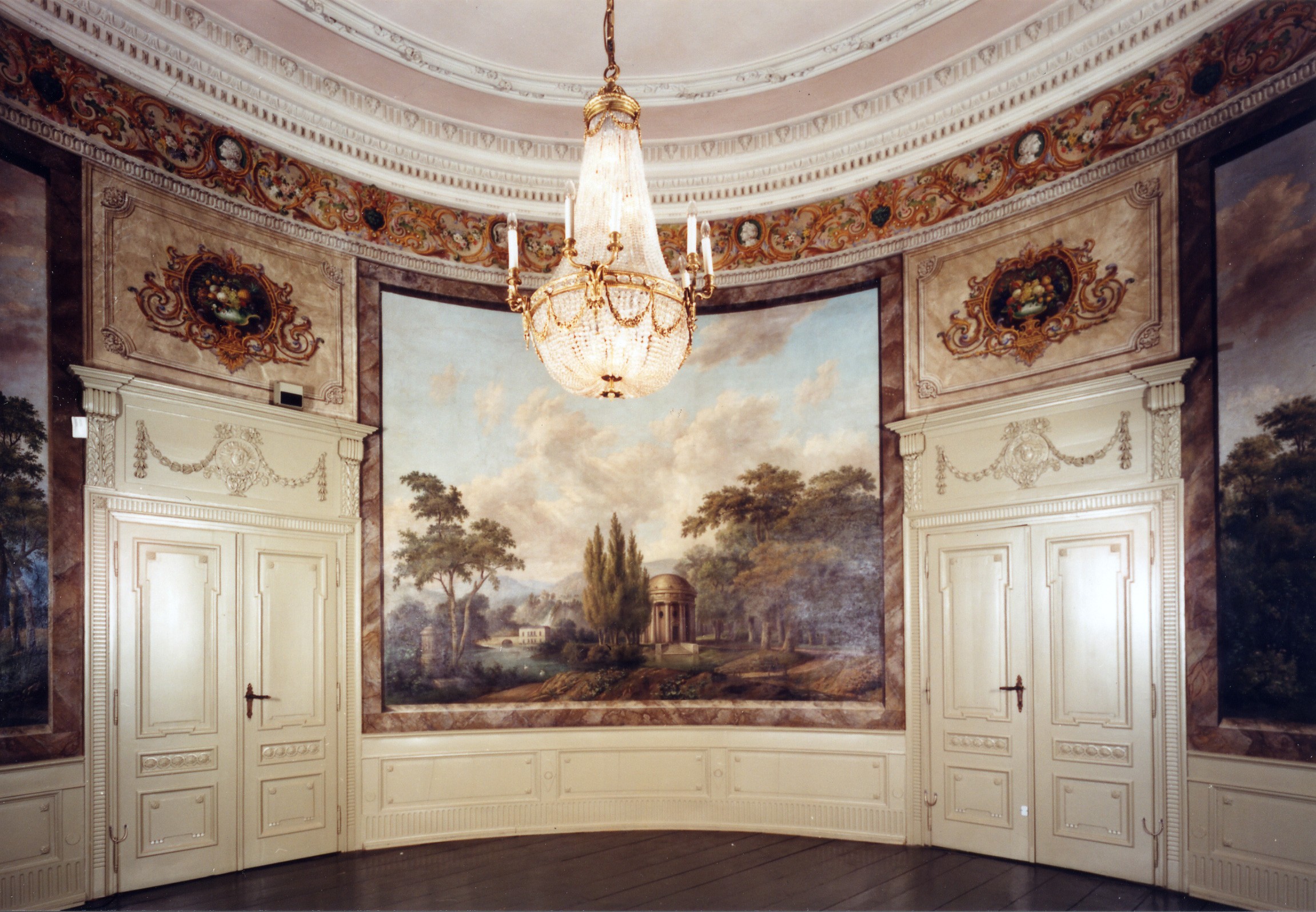 The representative highlight of the residence awaits the visitor on the first floor of the mansion: the Garden Room. The almost circular room extends over two storeys and is decorated with large classical landscape scenes. Tall Trumeau mirrors and console tables from the Empire period complete the stately impression of this room: it was Sophie Brügelmann who significantly influenced its present appearance in 1816. 