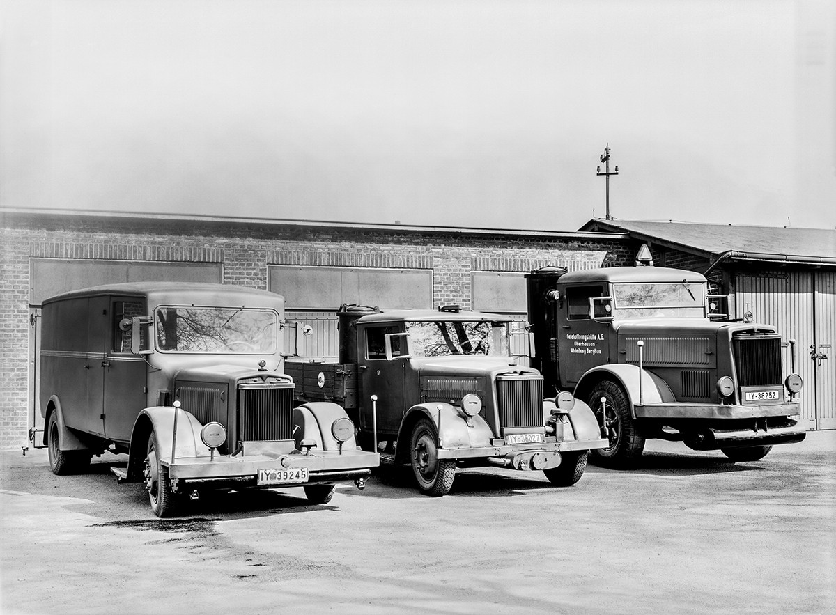 Historisches Schwarzweiß-Foto von drei Kraftfahrzeugen vor einer Wand. 