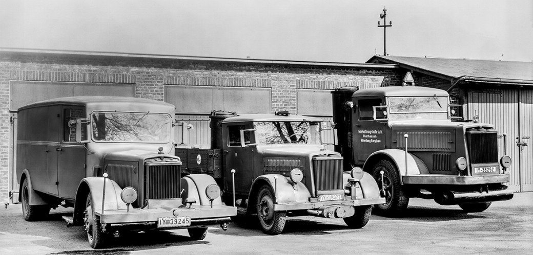 Historisches Schwarzweiß-Foto von drei Kraftfahrzeugen vor einer Wand. 