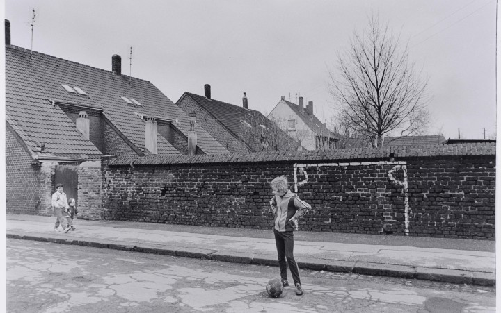 Schwarz-weiß-Fotografie einer Straßenszene, zeigt ein Kind mit Ball vor einer Wand mit aufgemaltem Tor
