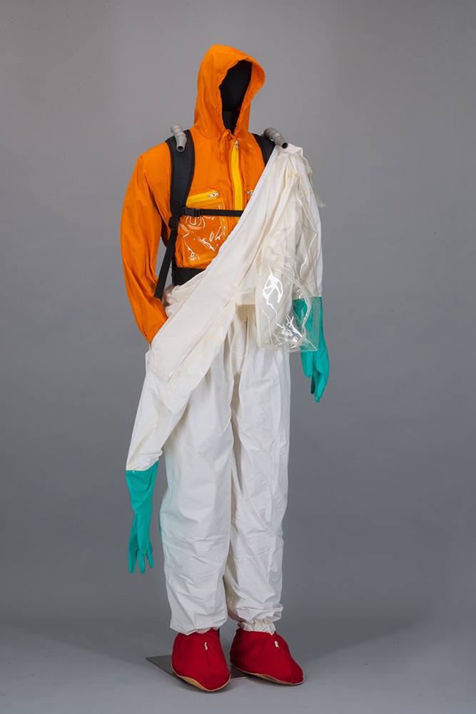 Eine Figurine trägt einen zweiteiligen Arbeitsschutzanzug: Über den inneren, orangenen Anzug ist zur Hälfte ein weißer Schutzanzug gestülpt. Die Puppe trägt dazu türkise Schutzhandschuhe und rote Schuhe
