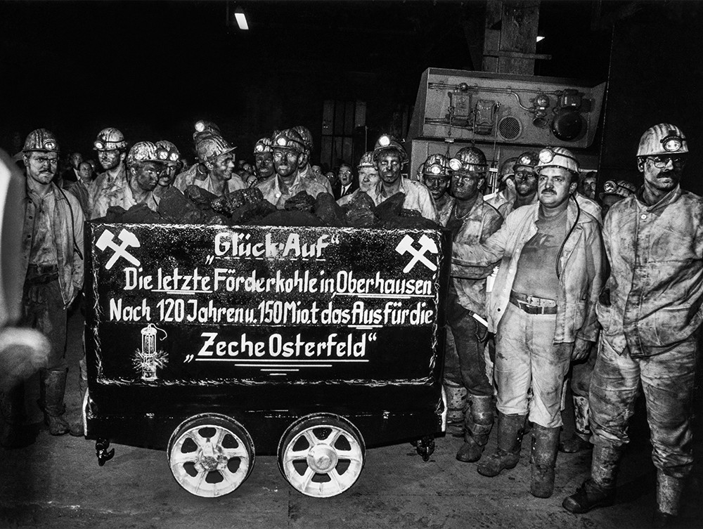 Schwarz-Weiß-Fotografie von eine Gruppe Bergleute, die Grubenhelme und verschmutze Arbeitskleidung tragen. Sie stehen um einen Kohle-Förderwagen herum, auf den geschrieben wurde "Glück auf. Die letzte Förderkohle in Oberhausen. Nach 120 Jahren und 150 Miollionen Tonnen das Aus für die Zeche Osterfeld".