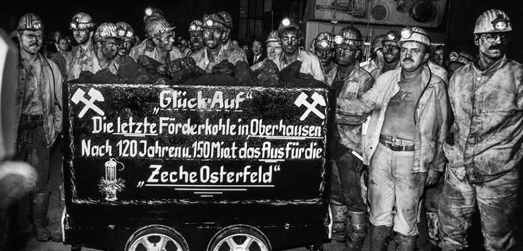 Schwarz-Weiß-Fotografie von eine Gruppe Bergleute, die Grubenhelme und verschmutze Arbeitskleidung tragen. Sie stehen um einen Kohle-Förderwagen herum, auf den geschrieben wurde "Glück auf. Die letzte Förderkohle in Oberhausen. Nach 120 Jahren und 150 Miollionen Tonnen das Aus für die Zeche Osterfeld".