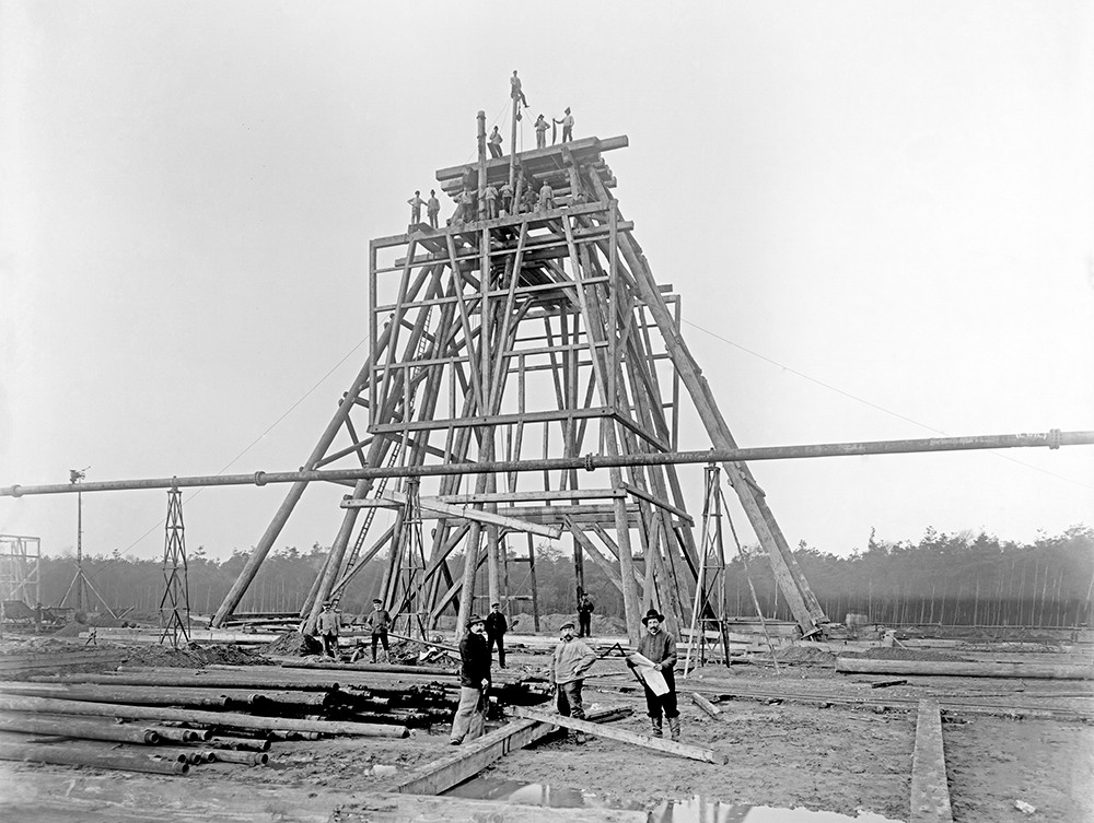 Die Schwarz-Weiß-Fotografie zeigt die Montage eines pyramidenförmigen Gerüstes. Auf der oberen Bauplattform des Gerüsts stehen 10 Monteure. Im Vordergrund stehen drei Männer neben Holzstämmen und Baumaterialien.