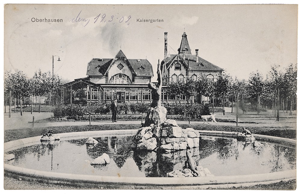 Vorderseite einer Postkarte zeigt Blick in einen Park mit Springbrunnen und Gebäude