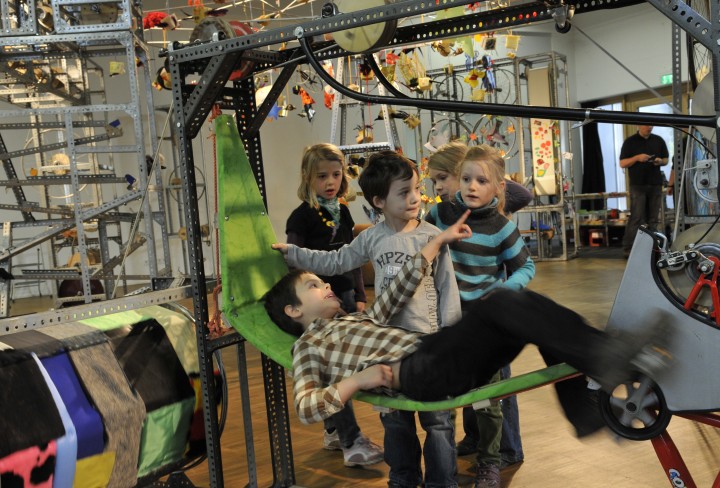 Eine Gruppe Kinder schaut zu, wie ein Kind einen Teil der Mitmach-Maschine mit einem Fahrradpedal in Bewegung setzt.
