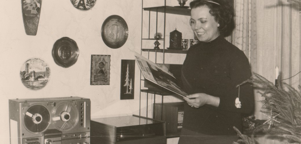 Schwarz weiß Fotografie einer Frau, die eine Schallplatte in den Händen hält und lacht. Sie steht neben einem Plattenspieler.