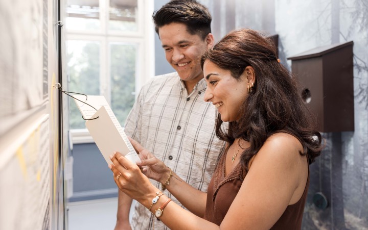 Zwei Personen stehen vor einer Wand mit einem aufgehängten Objekt, eine Person hält eine weiße, rechteckige Tafel in der Hand.