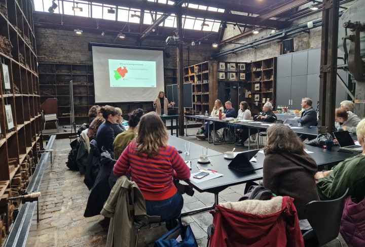 Projektteilnehmende sitzen an Tischen in U-Form und lauschen einem Vortrag.