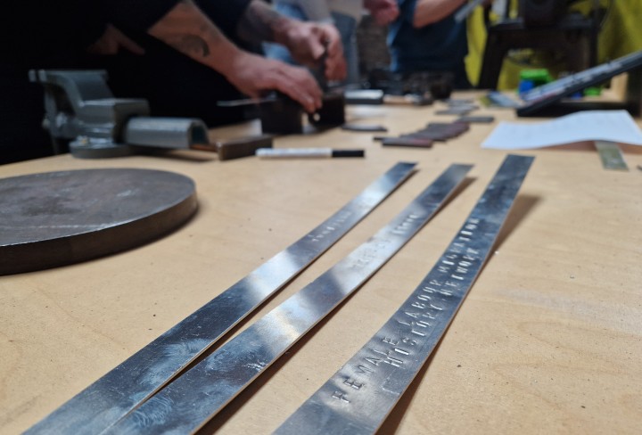 Drei lange, dünne Metallstücke mit eingeprägten Namen liegen auf einem Tisch. Im Hintergrund arbeiten Hände an weiteren Metallstücken.