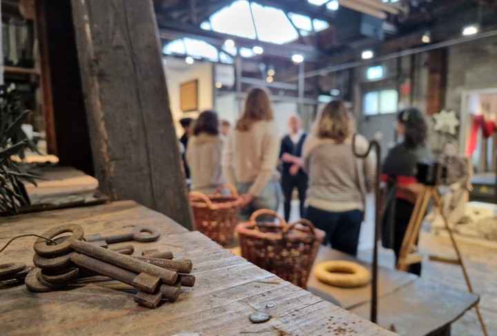 Blick Ã¼ber ein Regalbrett mit altem SchlÃ¼sselbund darauf auf eine Menschengruppe, die einem Vortrag lauscht.